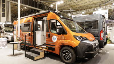 The Adria Twin Supreme 640 SGX is orange with a black roof, its sliding and rear doors are open.  There are two steps in front of the open sliding door to gain easy access.  Its outdoor table is fixed to the sliding door., 