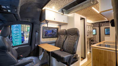 Knights Custom Conversion Prestige XL has wooden flooring with black upholstery. The cabinets are white and pale wood. The washroom door is open. The pop top roof is open.
