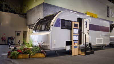 The Adria Alpina 623 UC Mississippi’s exterior has light grey walls with a patterned decal from its open door to the rear.  There are two wooden steps with an interactive display to the left.  There is an open locker. There is a floral display.