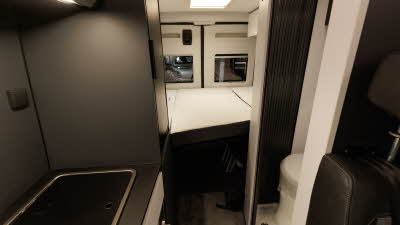 The Adria Twin Sports 600 SPB has black upholstery, the hob is to the left with the toilet opposite hidden bed a tambour door.  The fixed bed is at the rear.