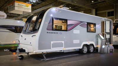 Bailey Phoenix 762 exterior’s grey and white with a large single front window.  The entrance door is open and there are fixed metal steps into its interior.  It is a twin axle. There is an interactive stand next to the steps.
