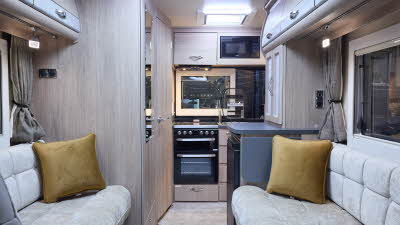 The Auto-Sleeper Nuevo has cream carpet, with pale wood cabinets with pale grey highlights. The upholstery is cream. There are two gold cushions. The kitchen is at the rear with an oven and the sink cover is open.