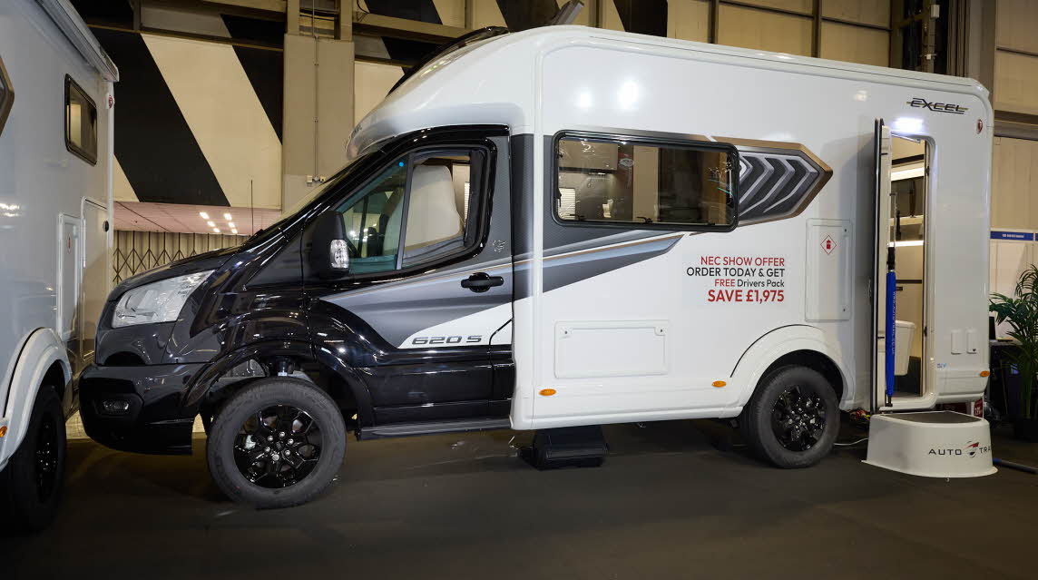 The Auto-Trail Excel 620S has a black cab and white body with grey decals.  There is a show offer on its side in red.  There is a white step by the opened habitation door which reveals the rear kitchen.