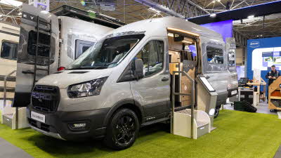 The Bailey Endurance E62 is silver, its sliding and rear doors are open.  There are two steps to gain easy access, there is an interactive stand next to the steps.