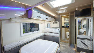 Mobilvetta K.Yacht 59-2’s interior has grey and white upholstery.  There are three skylights.  The rear internal door is open revealing the washroom.  Over the cupboards there is high level lighting.  To the left. there is a TV above a mirror.
