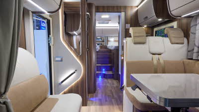 The Rimor Super Brig 95 Plus has a wooden floor.  The upholstery is cream, with gold accents.  The cabinets are all wooden and the interior door is a dark brown.  There are fixed rear beds.  The vehicle has a table in front of the travel seats.