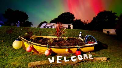 A yellow rowing boat filled with plants in front of a grassy bank with a caravan in the distance under a green and red sky