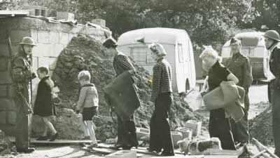 Citizens walk on duckboards past rubble with Home Guard soldiers looking on and caravans in the background