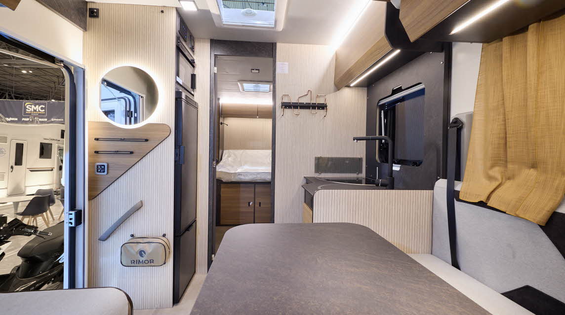 The Rimor Sarus 66 Plus has a cream floor.  The upholstery is cream, and it has gold curtains.  The cabinets are all wooden and the interior door is a dark brown.  There is a fixed rear bed.  The vehicle has a large table in front of the sofas.