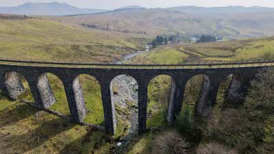 View from above looking across at Cwm Prysor Viaduct withh green fields and hills behind leading to mountains under the misty sky