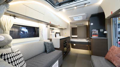 Adria Alpina Mississippi’s interior, it’s cream with a large panoramic window.  The upholstery is two tone grey.  The fixed bed is beyond the kitchen with the rear washroom’s door is open.