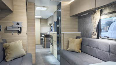 Interior of the Adria Altea Avon with grey sofas, cosy cushions and wood accents, illuminated by natural light through windows, offering a view outside.  Perfect for relaxation. and designed for comfort.