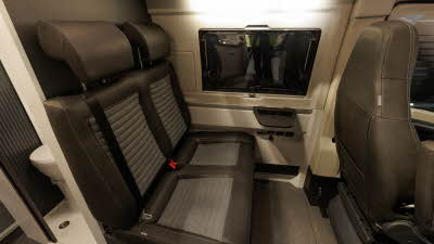 The Adria Twin Supreme 640 SGX has brown and grey upholstery.  There is a table for drinks.  To the bottom left is the toilet in the washroom with an open Tambour door.