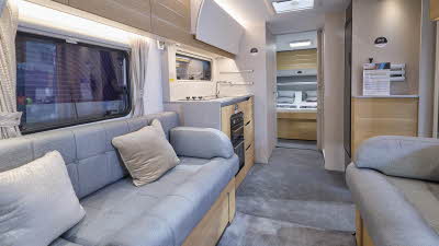 The interior of the Adria Adora 623 DP Tiber has cream walls.  The furniture is mid wood.  There are two sofas with grey upholstery and on one there are two cushions.  There is a large fixed bed at the rear.