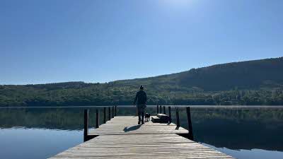 woman and dog walking on a pier over calm water reflecting the hills behind