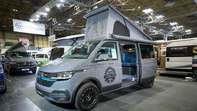 Vanworx Chesil’s exterior is grey with black decals.  It has a turquoise strip in the front grille.  The roof is fully extended.  Its sliding door is open revealing the lounge area.