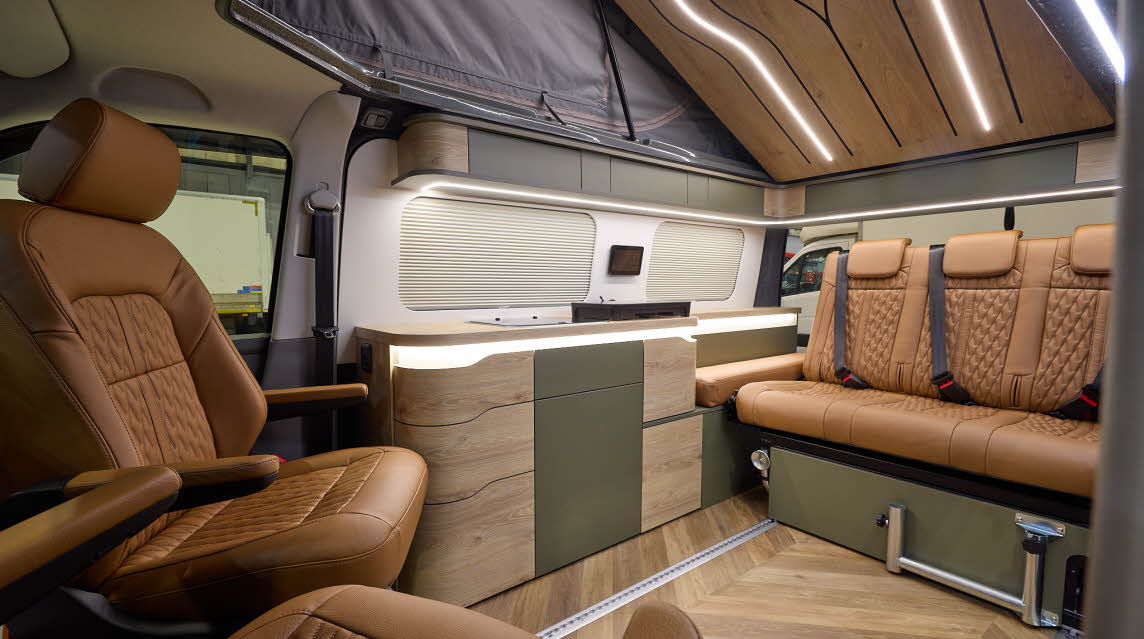 The Sherwood Campers Ltd Loxley Edition Conversion has wooden parquet-style flooring and its upholstery is brown leather.  Its furniture is wooden which is curved.  There are dark green inserts.  The rising roof is open, there's strip lighting.