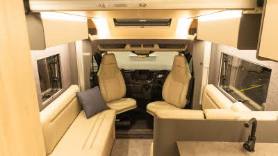 The Auto-Trail F-Line F60 has cream upholstery.  The two front seats are swivelled to the face the interior and are part of the lounge.  There is a grey cushion on the left hand sofa.  The overhead cabinets are cream with a brown surround.