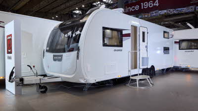 Elddis Avanté 868’s exterior is white with a small grey and dark red patterned decal. The entrance door is open and there is a large metal step to gain easy access.  The roof light is open and there is a TV aerial on the roof.