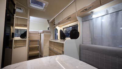 Eura Mobil Activa One 650 HS has a large white table and its upholstery is grey with white chequer pattern. The cabinets are cream and pale wood. There are three steps to the fixed rear bed.