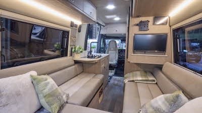 The RP Overland Rebel has wooden flooring. The upholstery is cream with a matching pattern. The cabinets are wooden. There is a television on the right hand wall. The passenger seat is facing into the vehicle. There is a u shaped lounge.