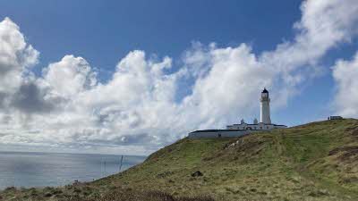 Lighthouse at the topof a grassy hill with a backdrop of blue sky and white clouds reflecting on the water below