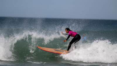 A surfer on a yellow board on a wave as it rolls over the board spraying water like a mist. The sky is pale blue as it meets the dark sea on the horizon