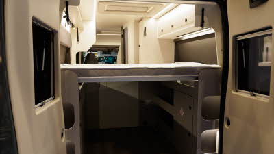 The Ace Motorhomes 1200 GL has a rear fixed bed which has a large storage area underneath.  There are three overhead lockers in white with square handles.  The rear doors are open.