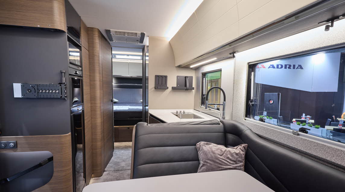 The Adria Supersonic’s floor has a grey carpet, with dark wood cabinets and cream curved overhead lockers.  The upholstery is dark leather.  There is a white table.  There is a fixed rear bed to the rear.