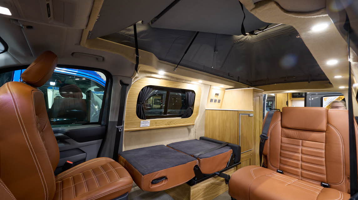 The Rolling Homes Camper Ltd Stanley has dark grey flooring.  The upholstery is brown leather.  The cabinets are wooden.  The rising roof is fully extended.  One of the passenger seats is folded down.