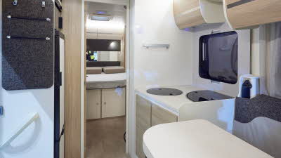 Rimor Evo 66 Plus has two tone grey upholstery.  The furniture is cream with pale wood doors.  The floor has beige vinyl.  Towards the rear is the bedroom with a fixed double bed.