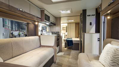 Interior of the Swift Conqueror Grande 645 with beige sofas, cosy cushions and beige carpets.   The large windows and three sky lights will let natural light flood in.  The door to the rear washroom, just beyond the bedroom, is open.