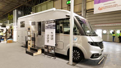 The Adria Supersonic 780 DL is silver.  There are two steps beside its habitation door.  There is a roller banner one side of the steps with an interaction stand on the other side.  Its skylights are open.