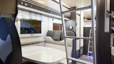 The Roller Team Zefiro 675 has grey carpet.  The upholstery is cream with dark brown highlights.  There is a cream table in front of the sofas.  The cabinets are cream with dark brown surrounds.  There are two ladders to gain access to beds.