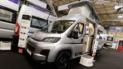 The Auto-Trail Expedition 67 is silver and has its rising roof extended.  Its sliding door is open and there is a step to enable easy access.  There is an information stand close to it.