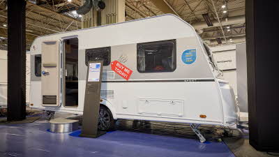Knaus Sport 460 EU is white with a grey decal.   The entrance door is open and there is a metal step to gain easy access.  There is a stand next to the steps