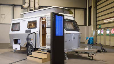 Eriba Touring 542 Legend’s exterior is silver and white.  It’s pop top roof is extended.  There are two wooden steps to gain easy access and there’s a large interactive screen to the right.