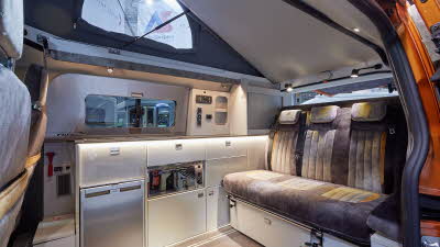 Auto-Sleeper Air has two tone grey embossed upholstery, with white furniture.  A fridge and mini grill is in the kitchen.  The rising roof is open and has a large semi-circle window in its canvas.