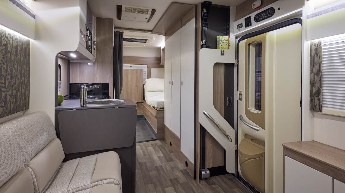 The Coachbuilt GB LIV Swift Escape 674 has a wooden floor, with cream cabinets.  The upholstery is cream and light brown.  There is fixed rear bed, close to the en suite washroom.