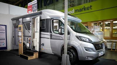 Adria Compact Supreme's exterior is silver, white and black, the entrance door is open and there are wooden steps to gain easy access.  There is an interactive information board by the side of the van.   