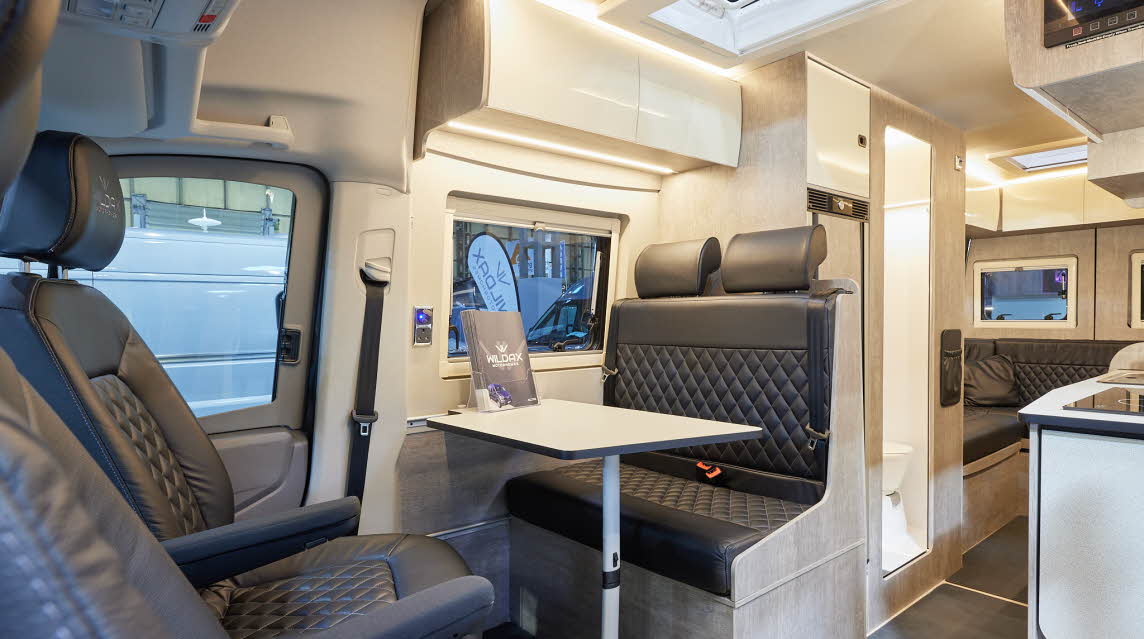 WildAx Altair has black upholstery; the panelling is wood.  The overhead lockers are cream.  The central washroom door is open.  There is a rear lounge.
