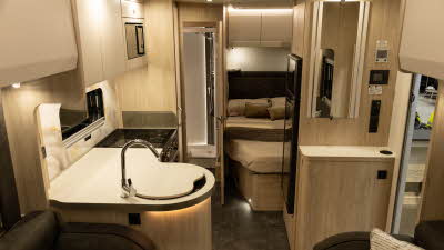 The Elddis Avalon 255 with XT Pack has dark brown upholstery complemented with pale wooden furniture and cream cabinet doors.  The kitchen is to the left and just beyond is a French transverse bed.  There is a rear washroom.