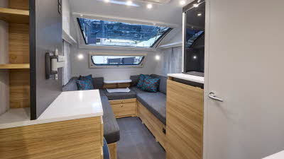Adria Action’s interior, it has a white door, walls and ceiling, with wooden furniture and white surfaces. The sofas have dark grey upholstery with blue/grey patterned cushions and the floor is dark grey. It has a large panoramic window.