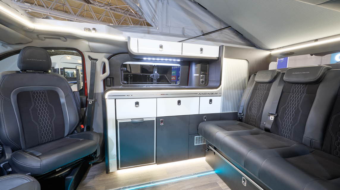 The Swift Monza has wooden flooring with black upholstery. The cabinets are white and dark blue. The pop top roof, along with its canvas, is open.