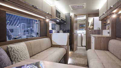 Interior of the Swift Conqueror 480 with beige sofas, cosy cushions and beige carpets.  There is a magazine at the front.  The large windows and two sky lights will let natural light flood in.  The door to the rear washroom is open.