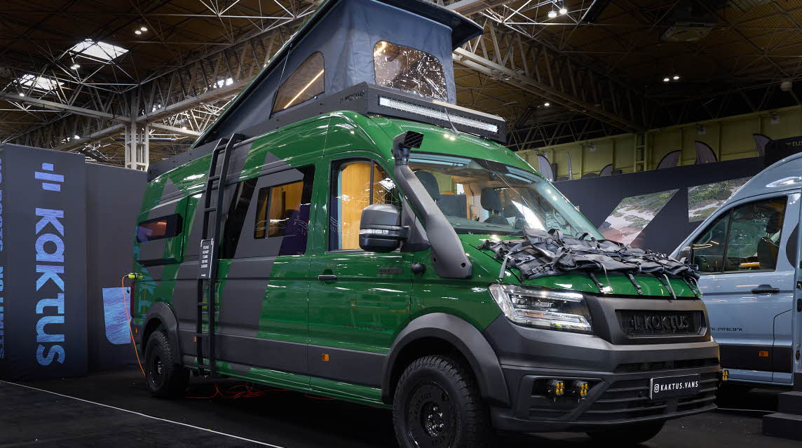 The Kaktus Adventure Vans Oasis x4 has a dark green and dark grey exterior with prominent decals.  There is an external ladder leading to the pop top roof which is fully extended.