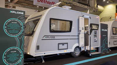 The Xplore XC DUO is white with grey sides and black decals.  There are two metal steps to gain easy access.  There is a large Xplore advertising banner next to the DUO. 