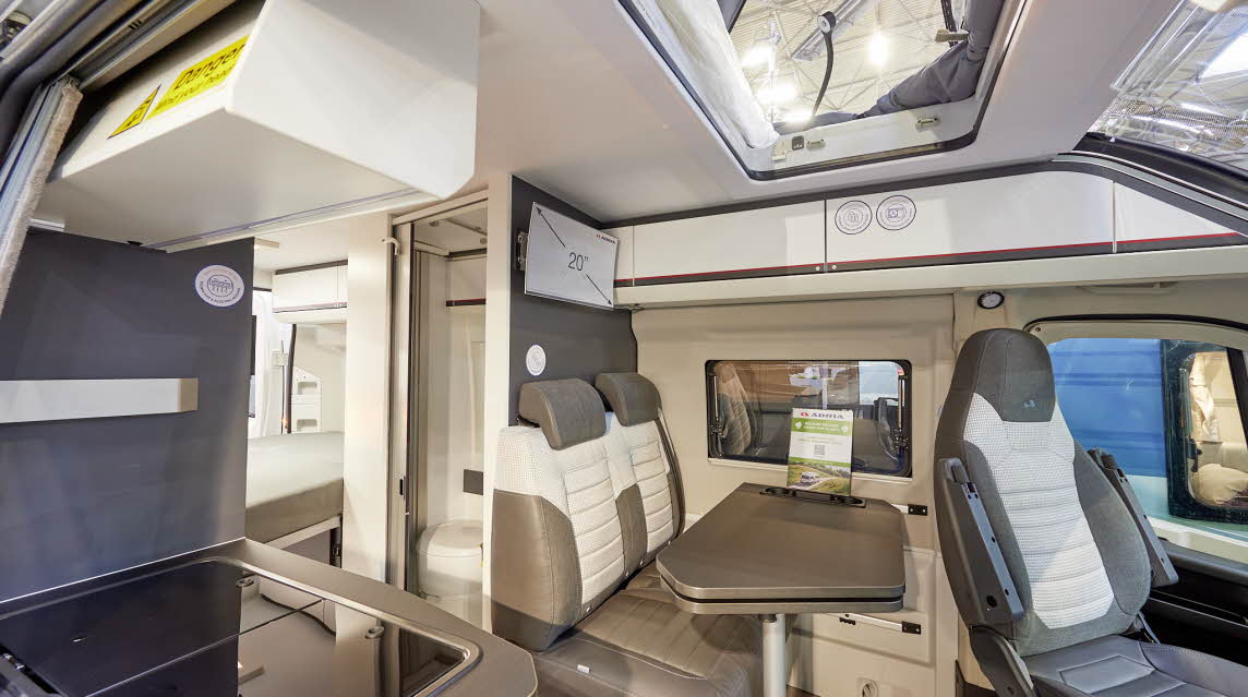 Adria Twin Sports 600 SPB interior, rear passenger seats in brown and cream, fixed table, swivelled driver’s seat which can double as a dining chair.  Also shown is a hob, access to the rising roof and the combined washroom and toilet.  