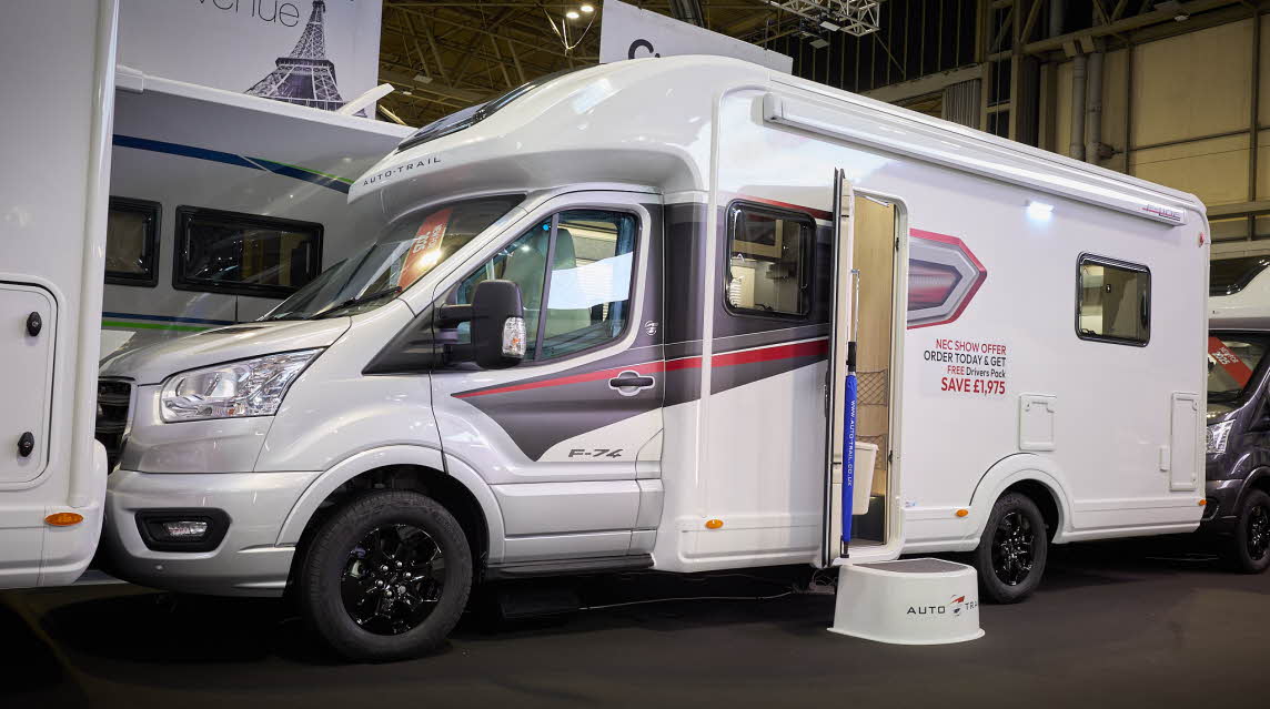 The Auto-Trail F-Line F74 has a white body with grey and red decals.  There is a show offer on its side in red.  There is a white step by the opened habitation door.  