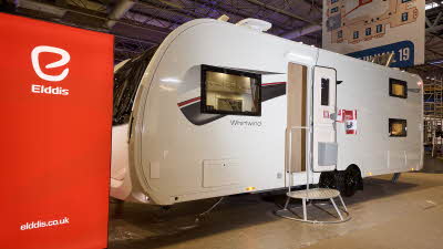 The Elddis Whirlwind Evolve 868 has a white exterior with a grey, red and black decal.  The entrance door is open and there is a metal step to gain easy access.  There is a large Elddis red banner with their website address next to it.